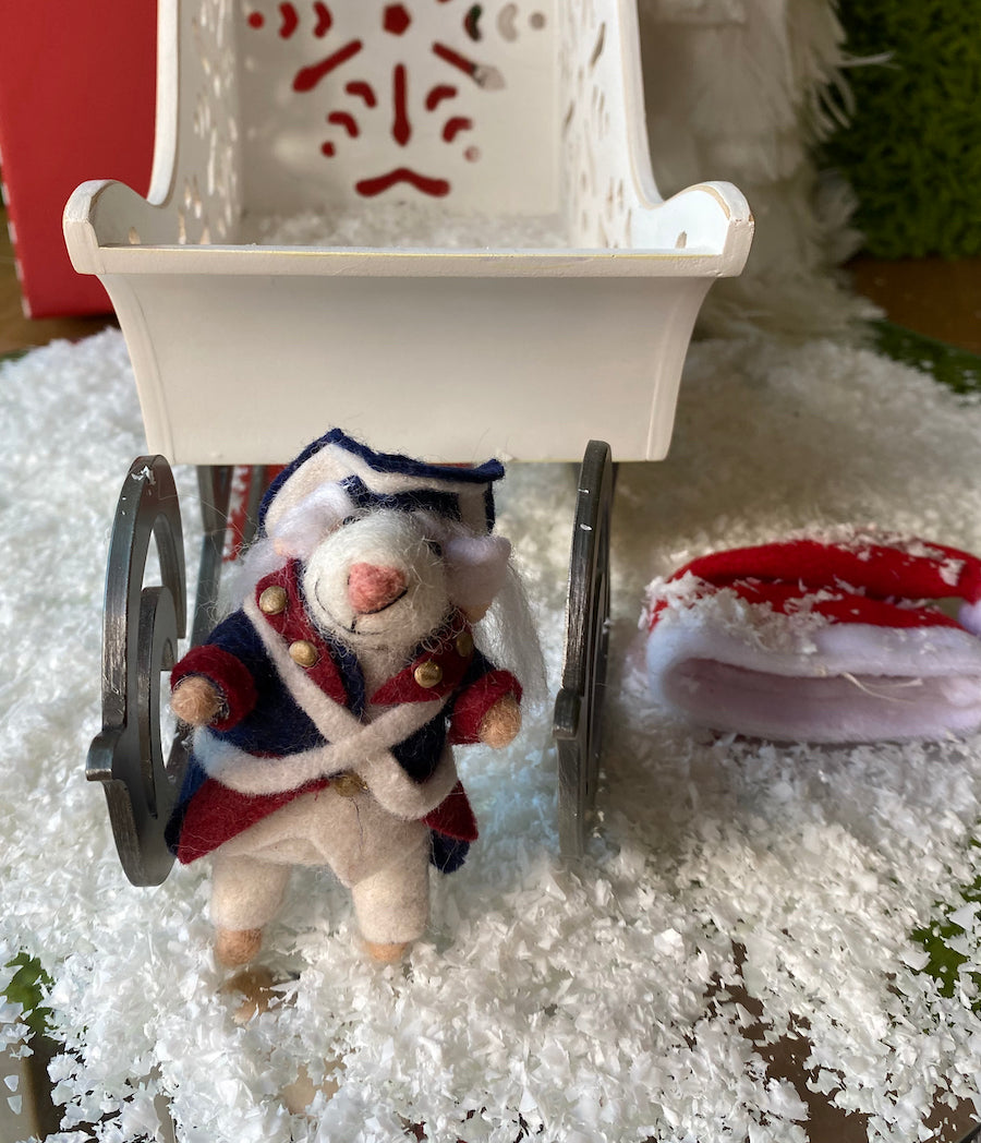 Brewmiceter Ornament Rosie Riveter Mouse standing in front of a white sleigh with snow on the ground and a Santa hat laying on the snow.